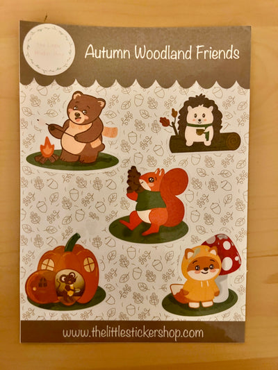 Sticker sheet with 5 cute character animals - a bear roasting a marshmallow over a campfire, a hedgehog drinking a hot drink on a log - a squirrel carrying a mountain of acorns, a fox in a raincoat, and a mouse in a pumpkin house.
