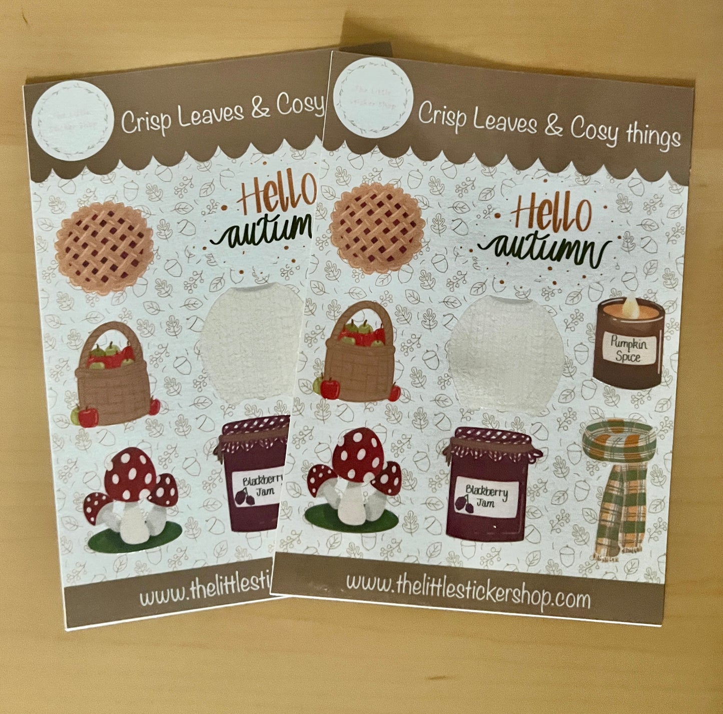 2 Autumn themed sticker sheets laid on a table, includes illustrations of a knitted jumper, scarf, pumpkin spice candle, blackberry jam, mushroom, apples in a basket, cherry pie and 'Hello Autumn' text. 