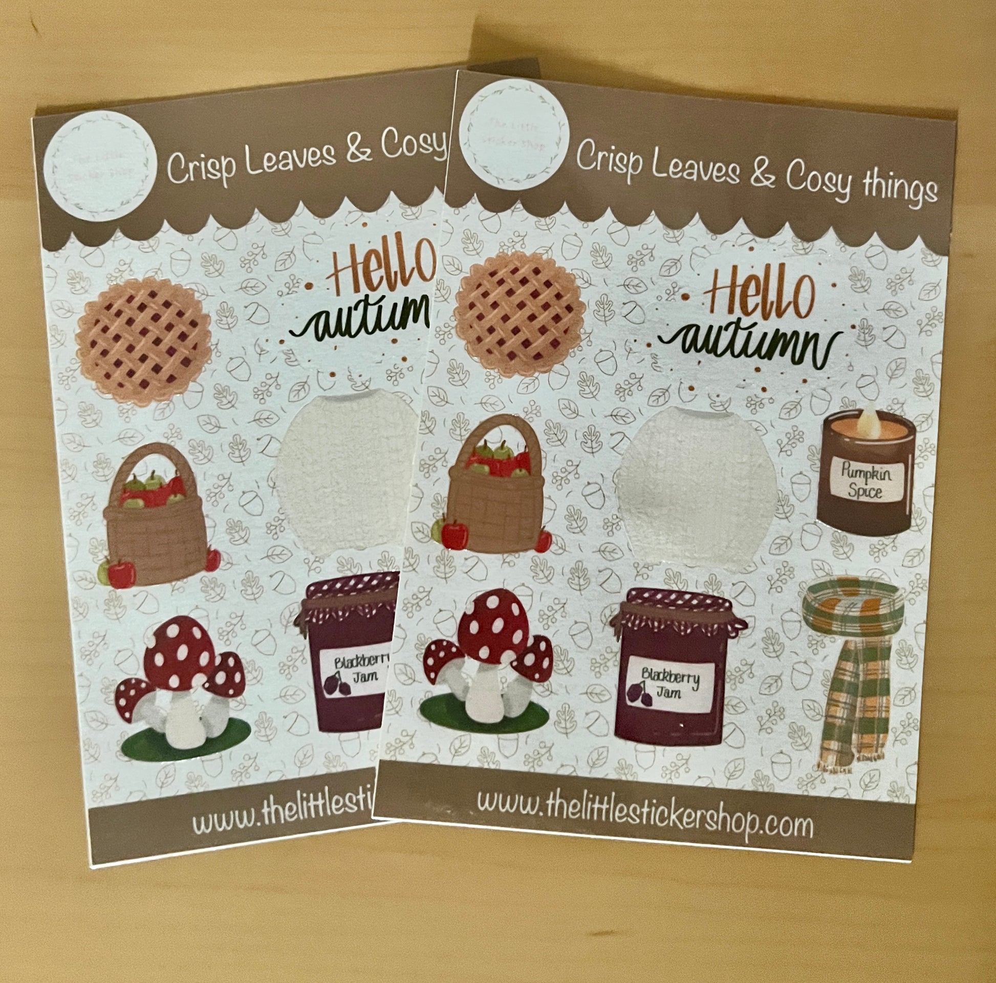 2 Autumn themed sticker sheets laid on a table, includes illustrations of a knitted jumper, scarf, pumpkin spice candle, blackberry jam, mushroom, apples in a basket, cherry pie and 'Hello Autumn' text. 
