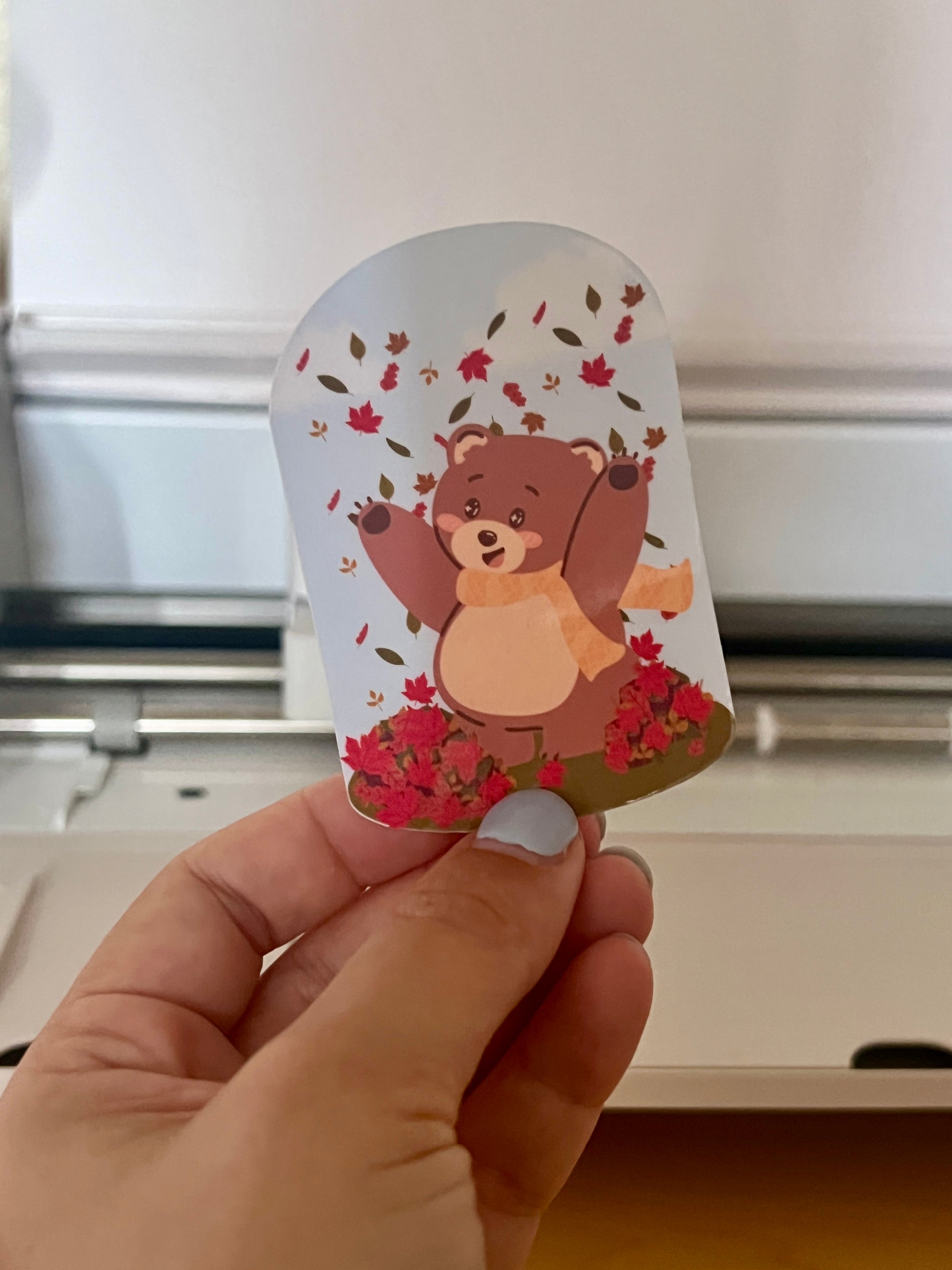 Die-cut sticker being held of a bear throwing leaves in the air. 