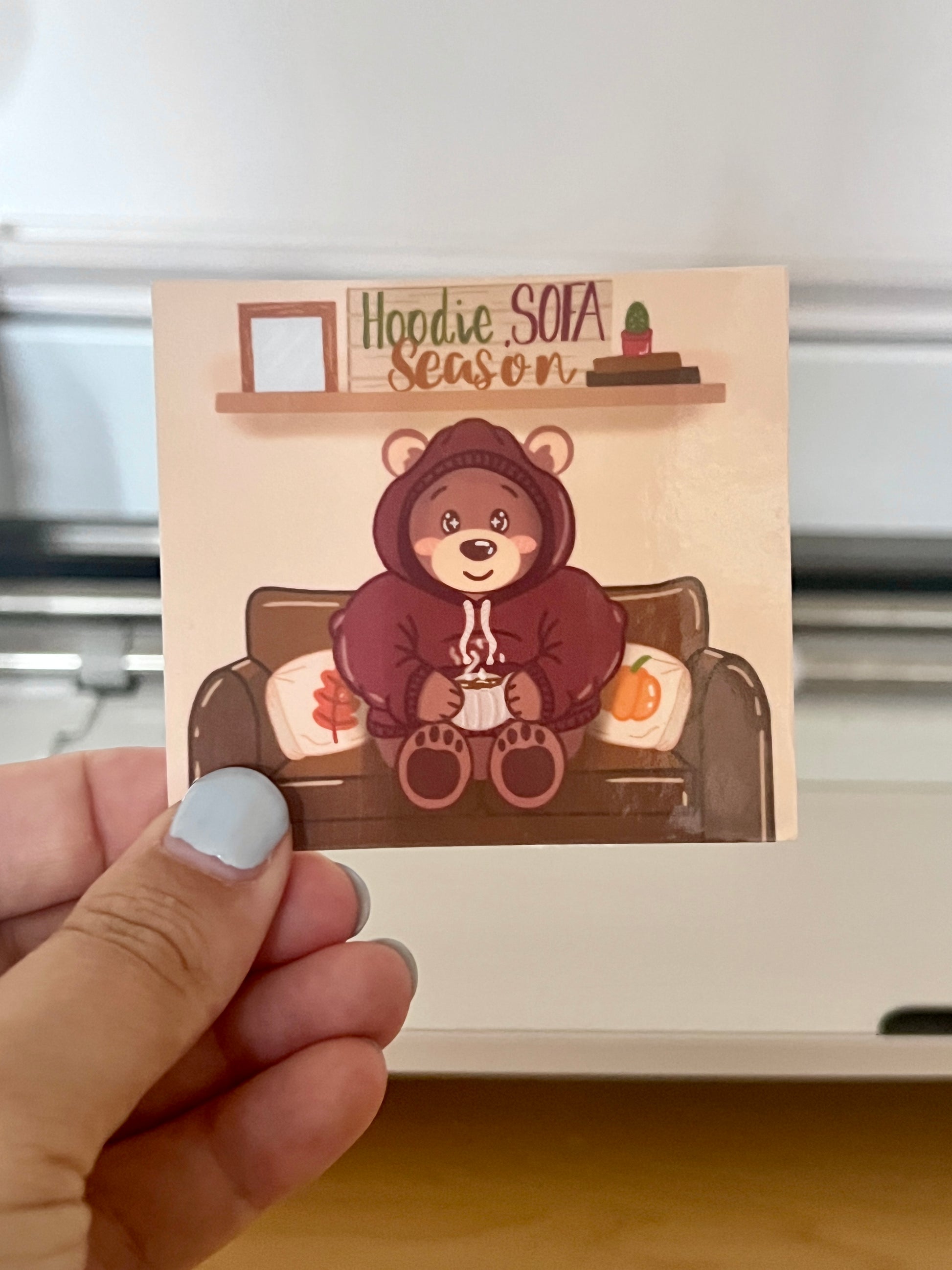 Die-cut sticker being held, of a bear in a hoodie drinking hot chocolate with text saying 'Hoodie, Sofa, Season'.