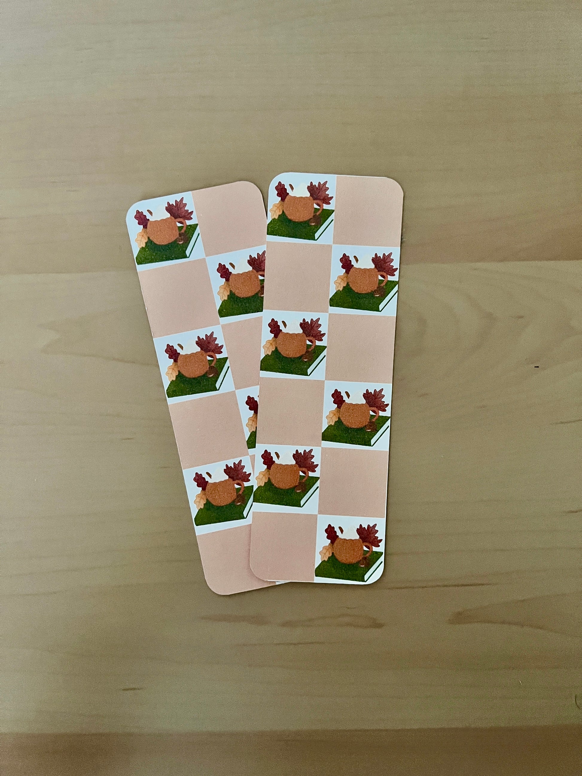 2 card bookmarks arranged on a wooden surface with illustrated pumpkin mug onto of a book with a chequered beige background. 